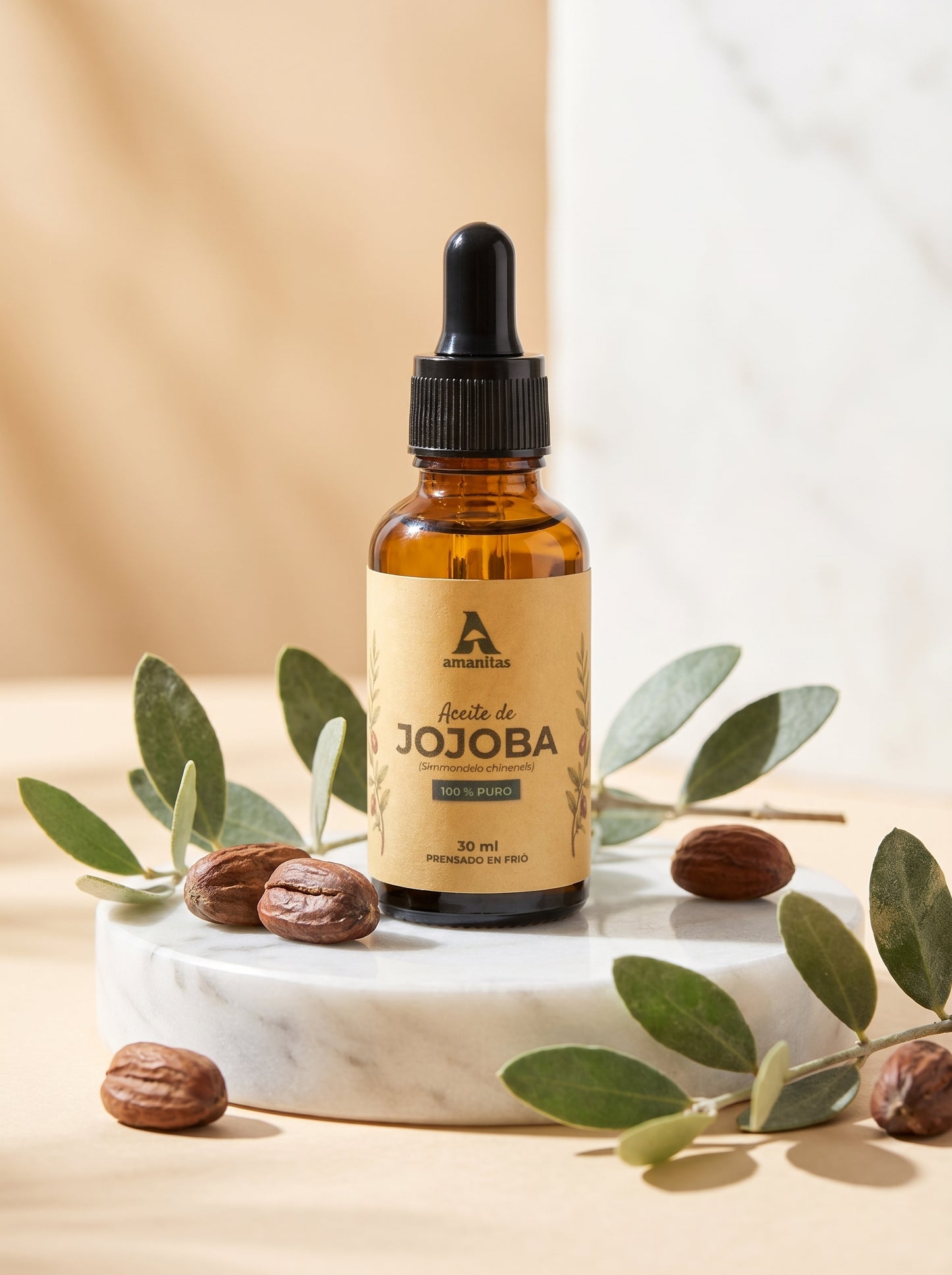 Frasco gotero de vidrio ámbar de Aceite de Jojoba 100% puro y prensado en frío de 30 ml, marca Amanitas. La etiqueta muestra una ilustración botánica de la planta de jojoba. El producto se exhibe sobre una base de mármol circular, rodeado de semillas de jojoba y hojas verdes frescas sobre un fondo neutro.