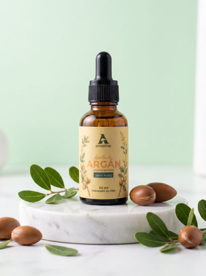 Frasco gotero de vidrio ámbar de Aceite de Argán 100% puro y prensado en frío de 30 ml, marca Amanitas. La etiqueta presenta una ilustración botánica en arte lineal de la especie Argania spinosa. El producto se exhibe sobre una base de mármol blanco rodeado de semillas de argán y hojas verdes.