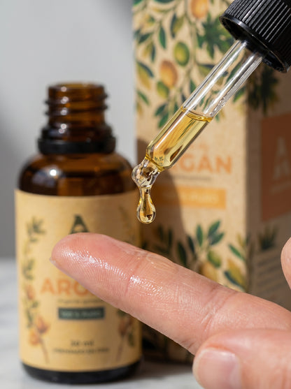 Primer plano de una gota de Aceite de Argán 100% puro cayendo desde un gotero hacia la yema de un dedo, mostrando su textura natural y fluida. Al fondo, se observa el frasco de vidrio ámbar de 30 ml de Amanitas con etiqueta botánica. Ideal para aplicación tópica en rostro y cabello.