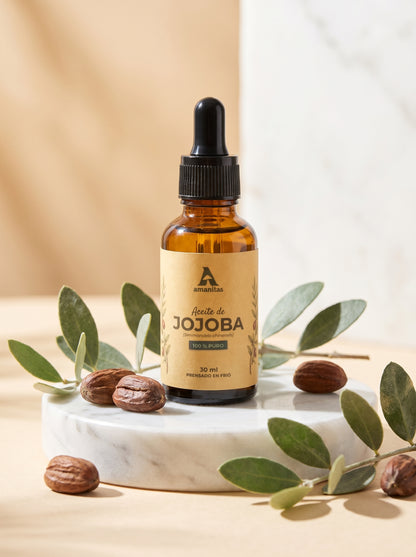 Frasco gotero de vidrio ámbar de Aceite de Jojoba 100% puro y prensado en frío de 30 ml, marca Amanitas. La etiqueta muestra una ilustración botánica de la planta de jojoba. El producto se exhibe sobre una base de mármol circular, rodeado de semillas de jojoba y hojas verdes frescas sobre un fondo neutro.