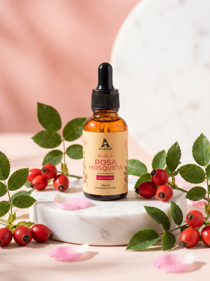 Frasco gotero de vidrio ámbar de 30 ml de Aceite de Rosa Mosqueta 100% puro y prensado en frío de Amanitas. El producto se exhibe sobre una base de mármol blanco, rodeado de frutos rojos y hojas frescas de rosa mosqueta, destacando su calidad artesanal y origen natural.
