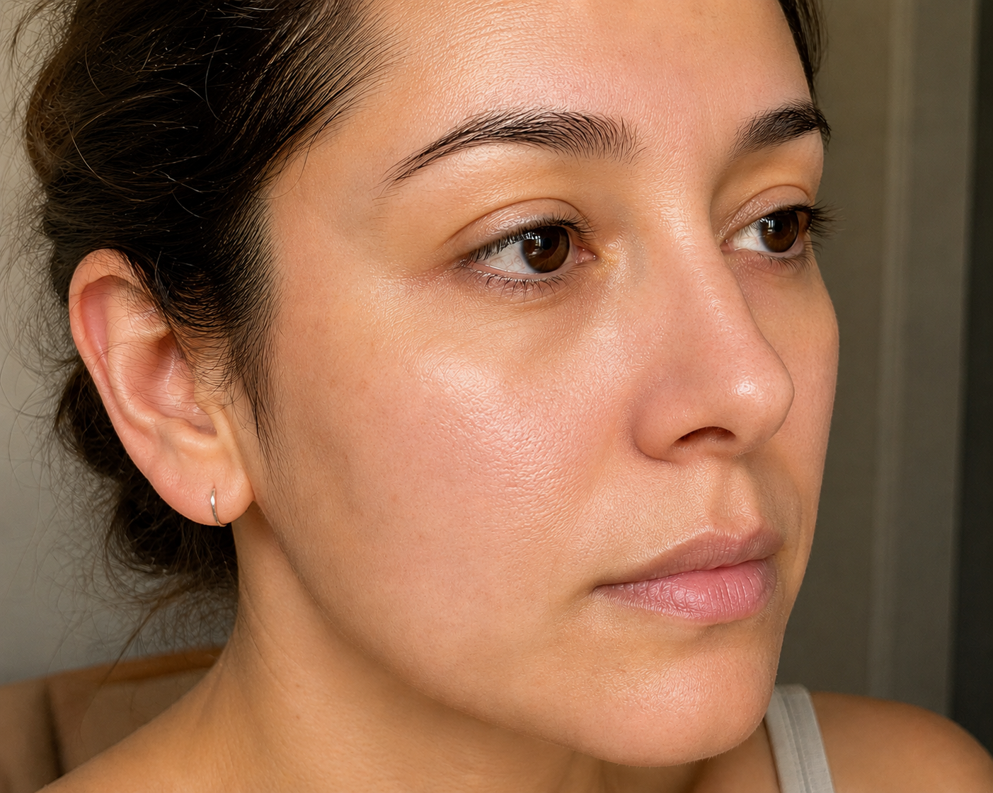 Retrato de una mujer joven con piel radiante y saludable y cabello rizado, con su mano cerca de su rostro, mostrando los resultados visibles de 'después' de usar los aceites botánicos naturales y puros de la marca Amanitas.pe, capturado contra una pared de crema texturizada.