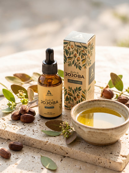 Presentación de Aceite de Jojoba 100% puro Amanitas en frasco gotero de 30 ml y caja, exhibidos sobre una base de piedra junto a un cuenco con aceite puro y semillas naturales. El diseño incluye ilustraciones botánicas de la planta de jojoba, destacando su calidad, origen natural y proceso de prensado en frío.